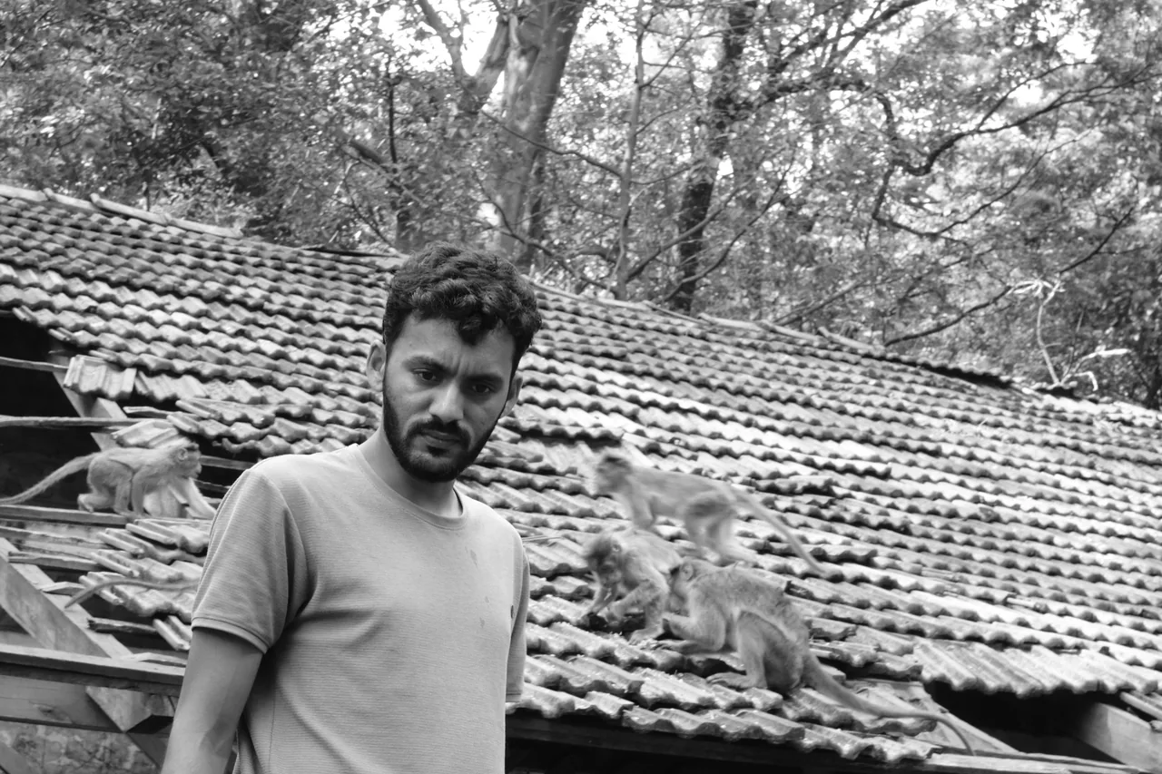 A man standing beside a tiled-roof building in a wooded area